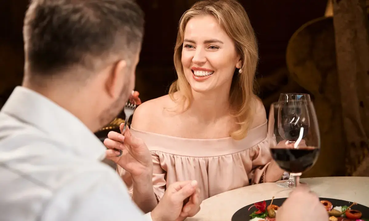 Coppia a cena, lei sorride mentre lui mangia. Forse stanno parlando di dove trovare sugar daddy per una vita di lusso.