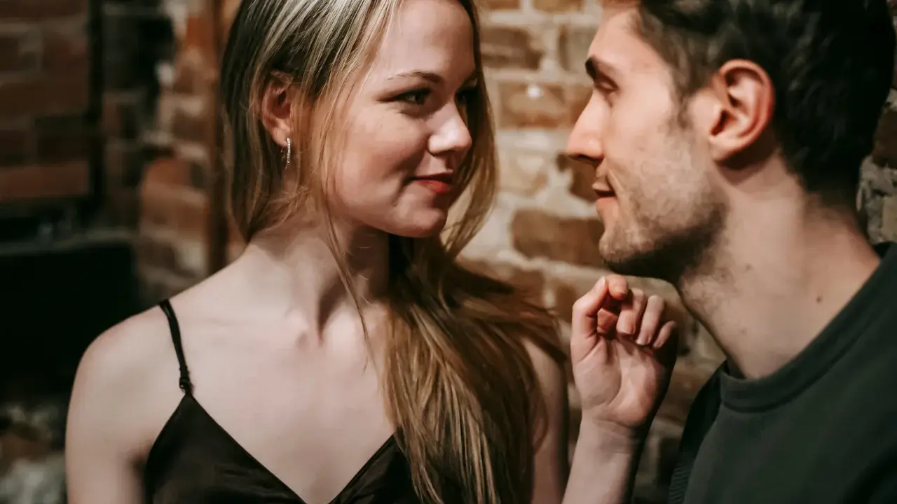 Un uomo e una donna si guardano intensamente. Lei ha lo sguardo di chi ti vuole, un sorriso accennato e i capelli biondi sciolti. Lui è vicino, con un'espressione concentrata.