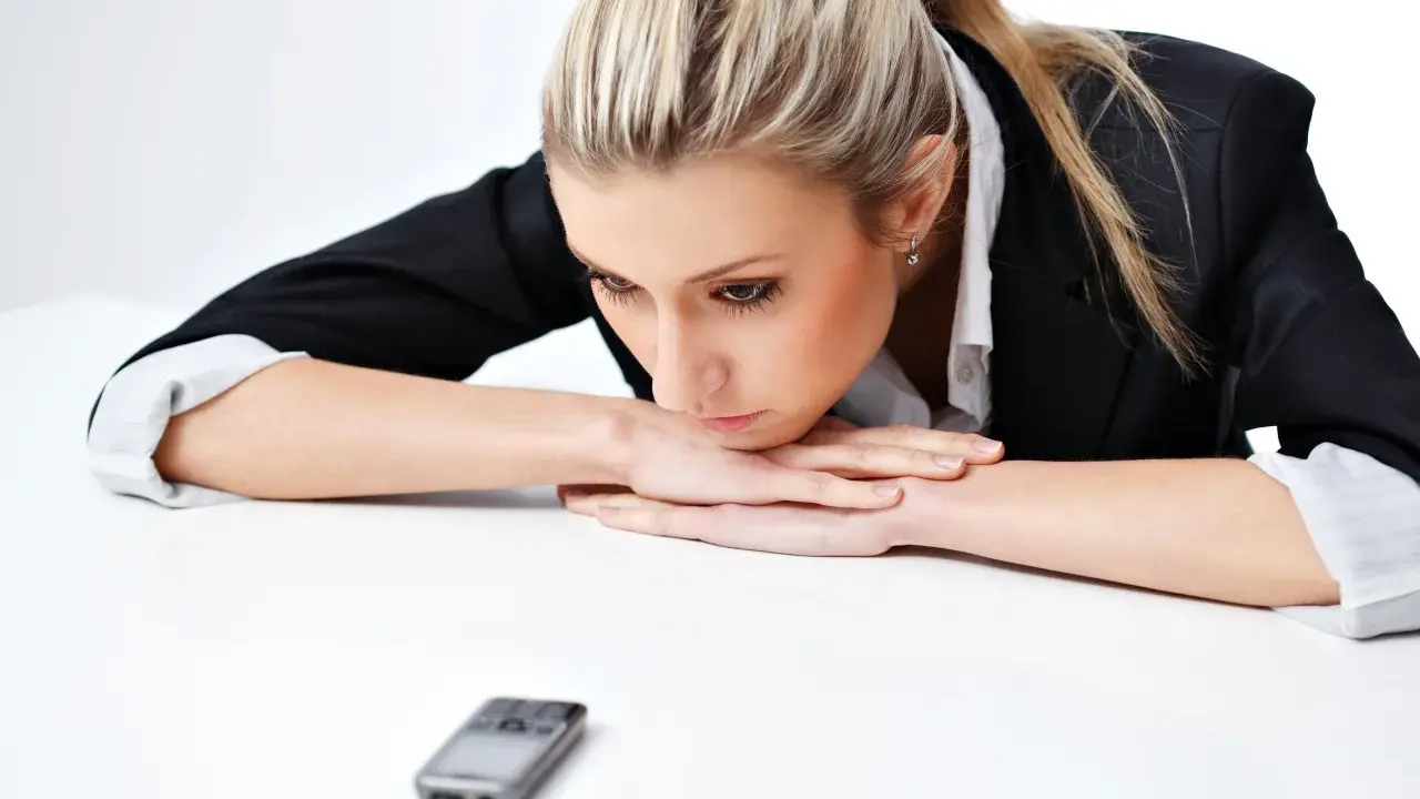 Donna bionda con giacca nera appoggiata sul tavolo, telefono in primo piano. Riflette su cosa scrivere ad un uomo che non ti cerca.
