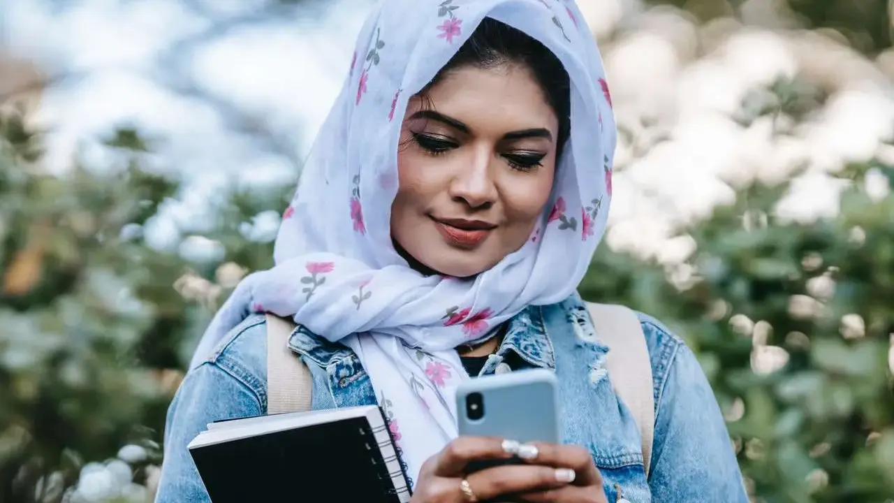 Ragazza con hijab fiorato guarda il telefono, indecisa se scrivere a un ragazzo o aspettare. Tiene un quaderno in mano.