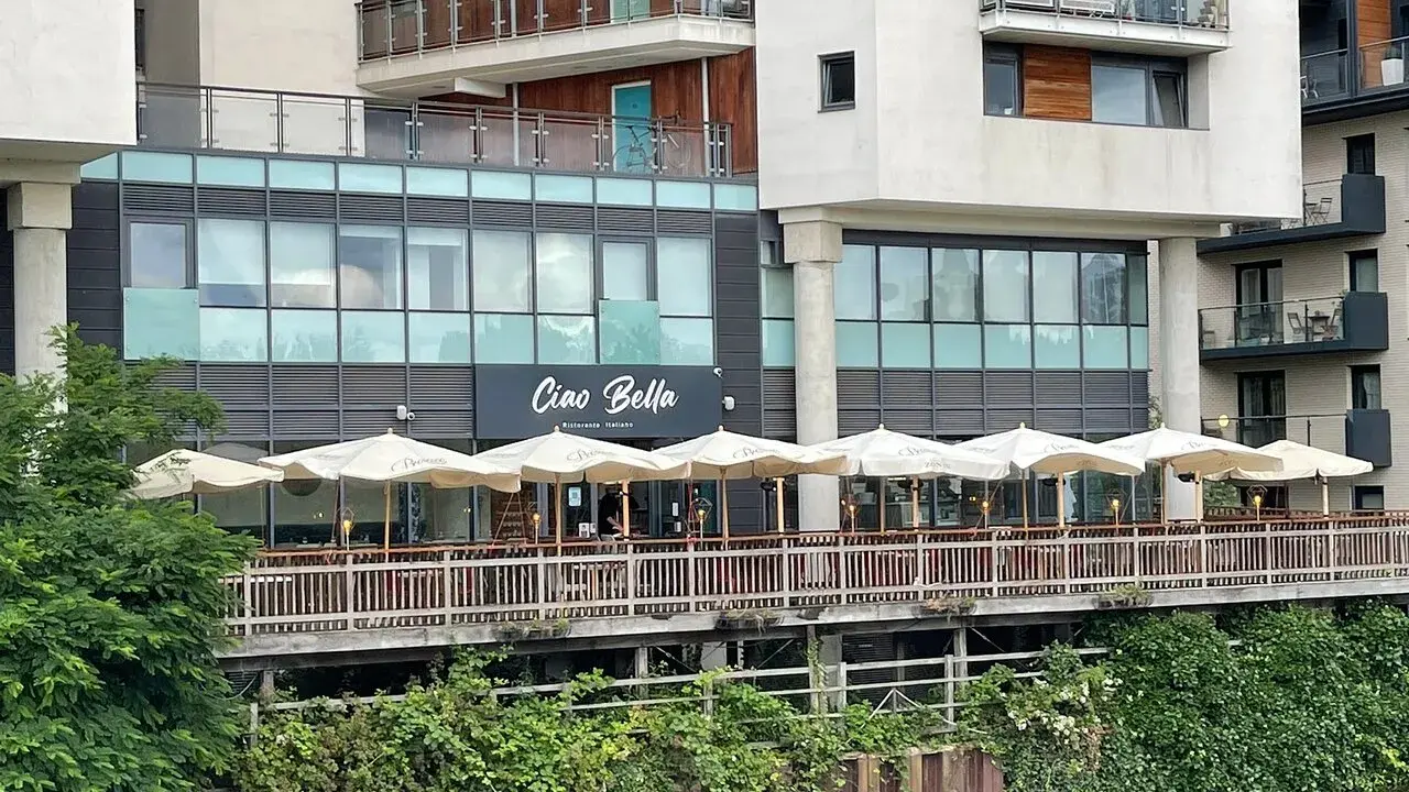 Ristorante "Ciao Bella" con tavoli all'aperto sotto ombrelloni bianchi, affacciato su un canale. Un complimento che invita a gustare la cucina italiana.