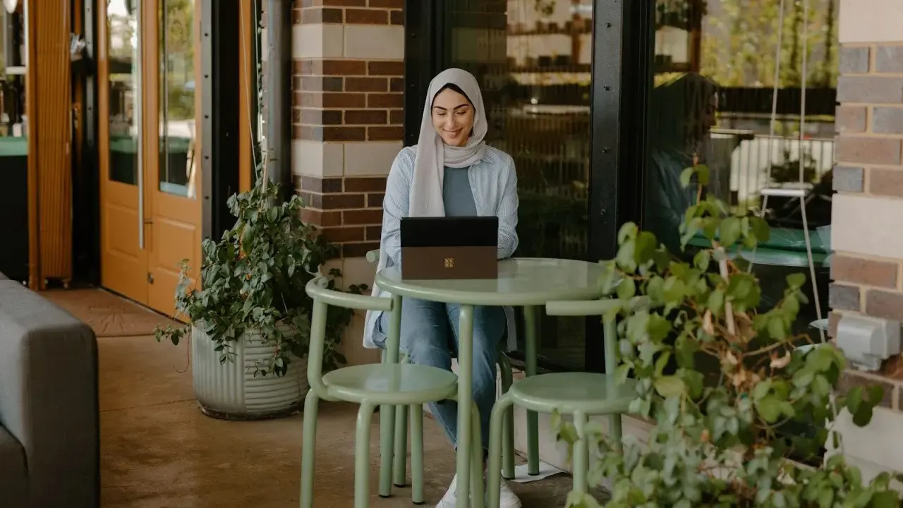 Donna con hijab sorride mentre lavora al laptop, seduta a un tavolino all'aperto. La scena suggerisce una **nirvam registrazione** di un momento di produttività serena.