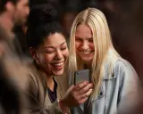 Due amiche ridono guardando il telefono, forse leggendo un divertente **meetic chat**.