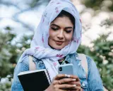 Ragazza con hijab fiorato guarda il telefono, indecisa se scrivere a un ragazzo o aspettare. Tiene un quaderno in mano.