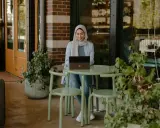 Donna con hijab sorride mentre lavora al laptop, seduta a un tavolino all'aperto. La scena suggerisce una **nirvam registrazione** di un momento di produttività serena.