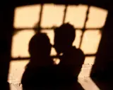 Ombre di una coppia che si abbraccia, illuminate da una finestra con grate, un'atmosfera intima che ricorda un incontro segreto in Italia.