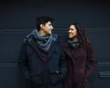 Un ragazzo e una ragazza si guardano negli occhi in silenzio, sorridendo. Indossano cappotti invernali e sciarpe.