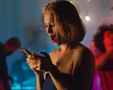 Sorpresa in chat: una ragazza con la bocca aperta guarda il suo telefono, mentre sullo sfondo si balla. Verità da fare in chat.