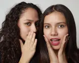 Due ragazze si sussurrano un segreto, mostrando un vivo interesse verso una persona. Una ha i capelli ricci e copre la bocca con la mano, l'altra ha i capelli lisci e un'espressione sorpresa.