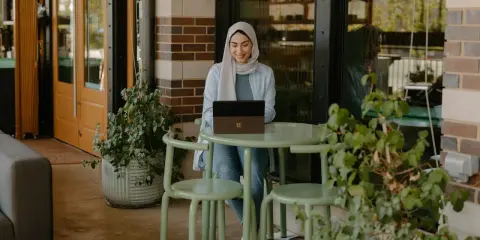 Donna con hijab sorride mentre lavora al laptop, seduta a un tavolino all'aperto. La scena suggerisce una **nirvam registrazione** di un momento di produttività serena.