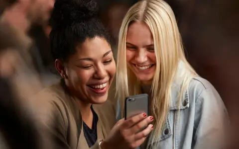 Due amiche ridono guardando il telefono, forse leggendo un divertente **meetic chat**.