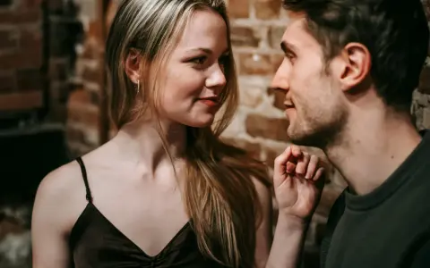 Un uomo e una donna si guardano intensamente. Lei ha lo sguardo di chi ti vuole, un sorriso accennato e i capelli biondi sciolti. Lui è vicino, con un'espressione concentrata.