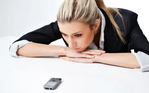 Donna bionda con giacca nera appoggiata sul tavolo, telefono in primo piano. Riflette su cosa scrivere ad un uomo che non ti cerca.