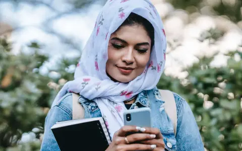 Ragazza con hijab fiorato guarda il telefono, indecisa se scrivere a un ragazzo o aspettare. Tiene un quaderno in mano.