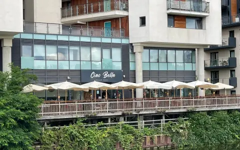 Ristorante "Ciao Bella" con tavoli all'aperto sotto ombrelloni bianchi, affacciato su un canale. Un complimento che invita a gustare la cucina italiana.