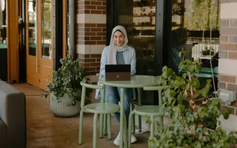 Donna con hijab sorride mentre lavora al laptop, seduta a un tavolino all'aperto. La scena suggerisce una **nirvam registrazione** di un momento di produttività serena.