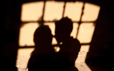 Ombre di una coppia che si abbraccia, illuminate da una finestra con grate, un'atmosfera intima che ricorda un incontro segreto in Italia.