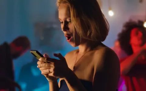 Sorpresa in chat: una ragazza con la bocca aperta guarda il suo telefono, mentre sullo sfondo si balla. Verità da fare in chat.