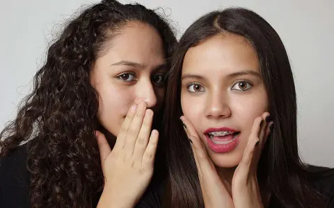 Due ragazze si sussurrano un segreto, mostrando un vivo interesse verso una persona. Una ha i capelli ricci e copre la bocca con la mano, l'altra ha i capelli lisci e un'espressione sorpresa.