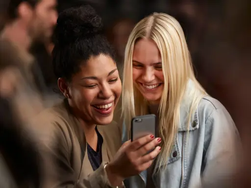 Due amiche ridono guardando il telefono, forse leggendo un divertente **meetic chat**.