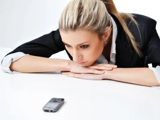 Donna bionda con giacca nera appoggiata sul tavolo, telefono in primo piano. Riflette su cosa scrivere ad un uomo che non ti cerca.