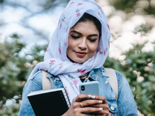Ragazza con hijab fiorato guarda il telefono, indecisa se scrivere a un ragazzo o aspettare. Tiene un quaderno in mano.