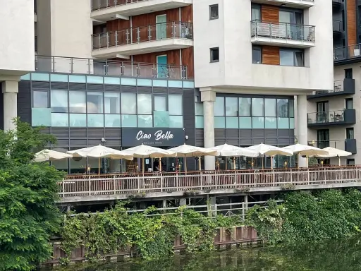 Ristorante "Ciao Bella" con tavoli all'aperto sotto ombrelloni bianchi, affacciato su un canale. Un complimento che invita a gustare la cucina italiana.