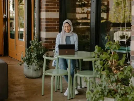 Donna con hijab sorride mentre lavora al laptop, seduta a un tavolino all'aperto. La scena suggerisce una **nirvam registrazione** di un momento di produttività serena.
