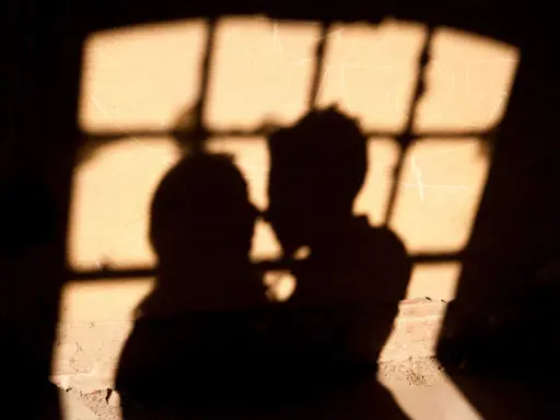 Ombre di una coppia che si abbraccia, illuminate da una finestra con grate, un'atmosfera intima che ricorda un incontro segreto in Italia.
