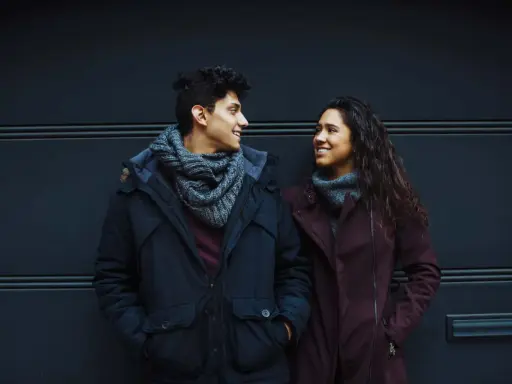 Un ragazzo e una ragazza si guardano negli occhi in silenzio, sorridendo. Indossano cappotti invernali e sciarpe.