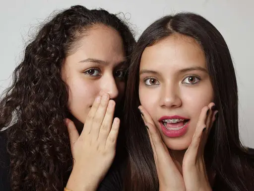 Due ragazze si sussurrano un segreto, mostrando un vivo interesse verso una persona. Una ha i capelli ricci e copre la bocca con la mano, l'altra ha i capelli lisci e un'espressione sorpresa.