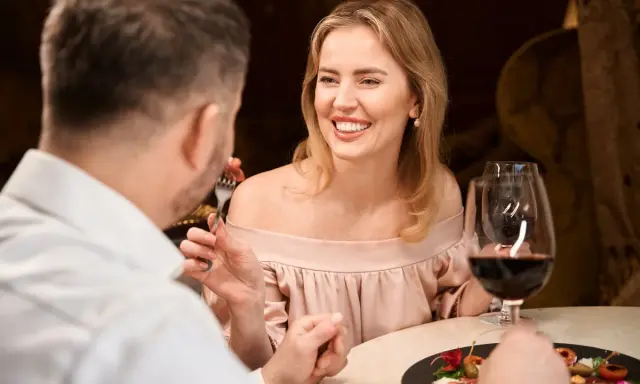 Coppia a cena, lei sorride mentre lui mangia. Forse stanno parlando di dove trovare sugar daddy per una vita di lusso.