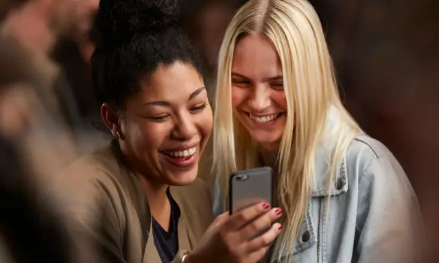 Due amiche ridono guardando il telefono, forse leggendo un divertente **meetic chat**.