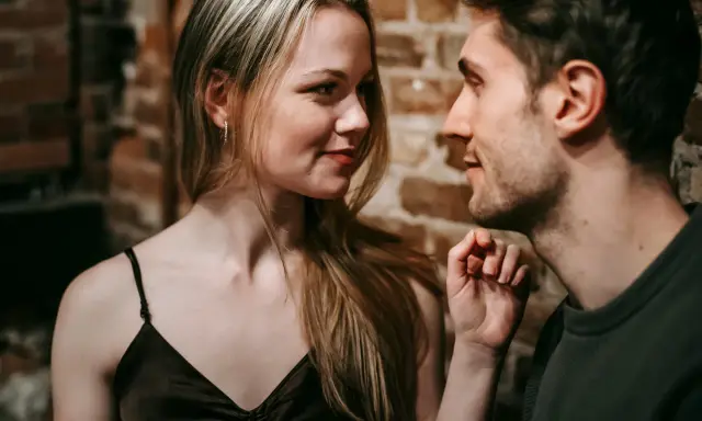 Un uomo e una donna si guardano intensamente. Lei ha lo sguardo di chi ti vuole, un sorriso accennato e i capelli biondi sciolti. Lui è vicino, con un'espressione concentrata.
