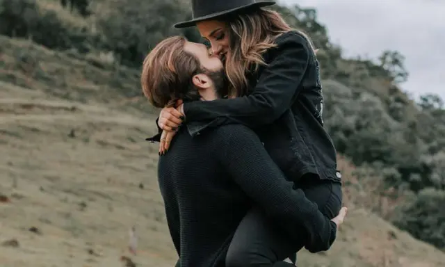 Coppia in un abbraccio appassionato all'aperto. Lui la solleva, lei indossa un cappello. Lui risponde ai messaggi ma non scrive per primo, ma in questo momento è tutto ciò che conta.