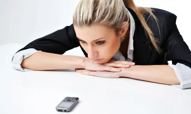 Donna bionda con giacca nera appoggiata sul tavolo, telefono in primo piano. Riflette su cosa scrivere ad un uomo che non ti cerca.