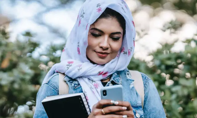 Ragazza con hijab fiorato guarda il telefono, indecisa se scrivere a un ragazzo o aspettare. Tiene un quaderno in mano.