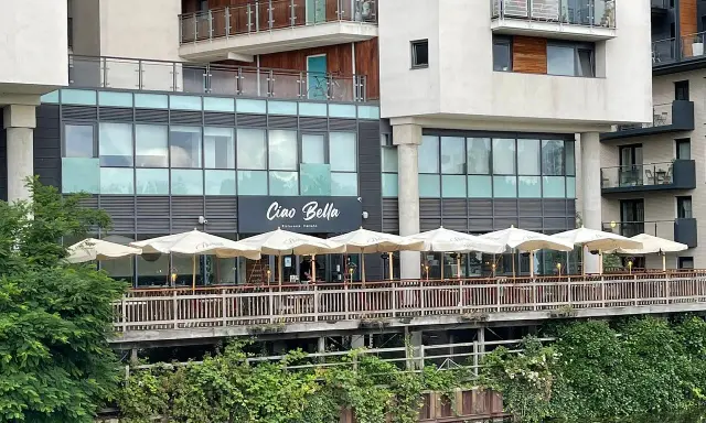 Ristorante "Ciao Bella" con tavoli all'aperto sotto ombrelloni bianchi, affacciato su un canale. Un complimento che invita a gustare la cucina italiana.