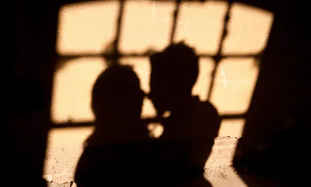 Ombre di una coppia che si abbraccia, illuminate da una finestra con grate, un'atmosfera intima che ricorda un incontro segreto in Italia.