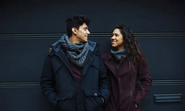 Un ragazzo e una ragazza si guardano negli occhi in silenzio, sorridendo. Indossano cappotti invernali e sciarpe.