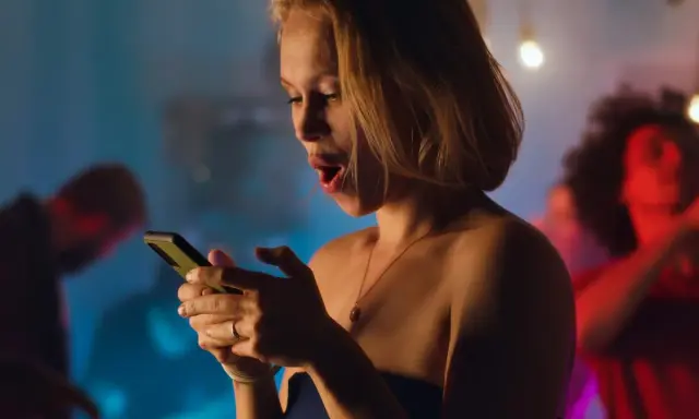 Sorpresa in chat: una ragazza con la bocca aperta guarda il suo telefono, mentre sullo sfondo si balla. Verità da fare in chat.