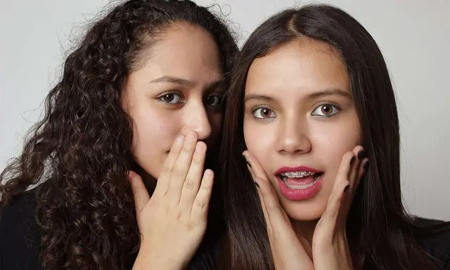 Due ragazze si sussurrano un segreto, mostrando un vivo interesse verso una persona. Una ha i capelli ricci e copre la bocca con la mano, l'altra ha i capelli lisci e un'espressione sorpresa.