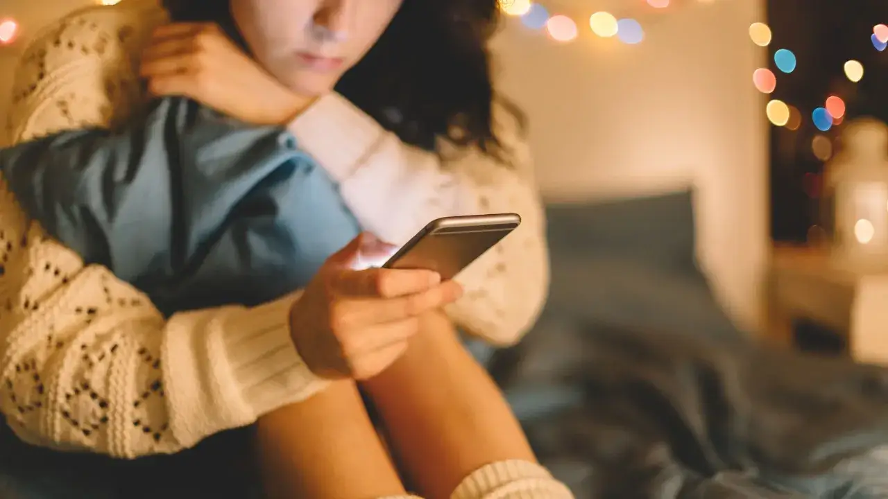 Ragazza con maglione bianco e calzini alti, abbraccia un cuscino blu, immersa nel suo smartphone. Luci soffuse e colorate creano un'atmosfera intima, perfetta per una chiacchierata che ti passa.