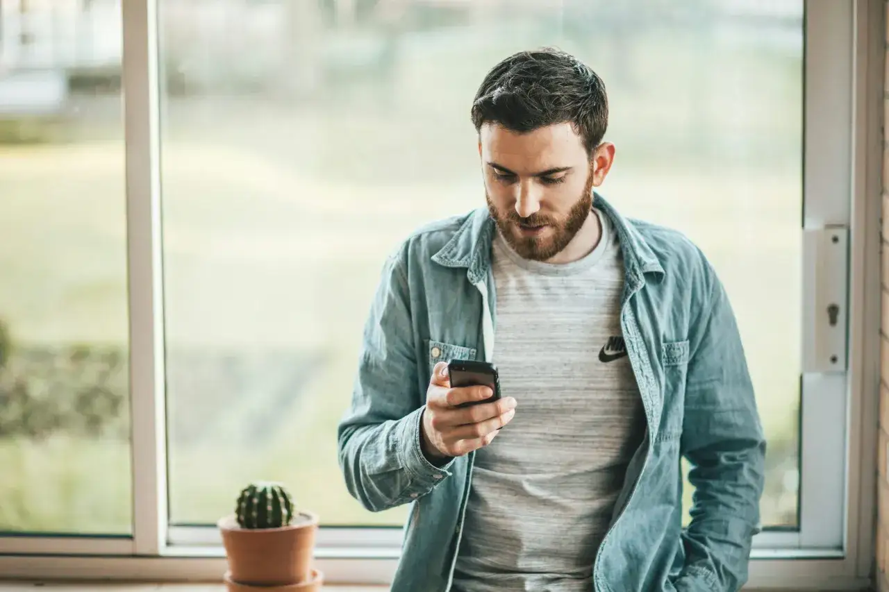 Uomo con barba guarda il telefono, pensando cosa pensa un uomo se non ti fai sentire. Vicino a una finestra con un cactus.