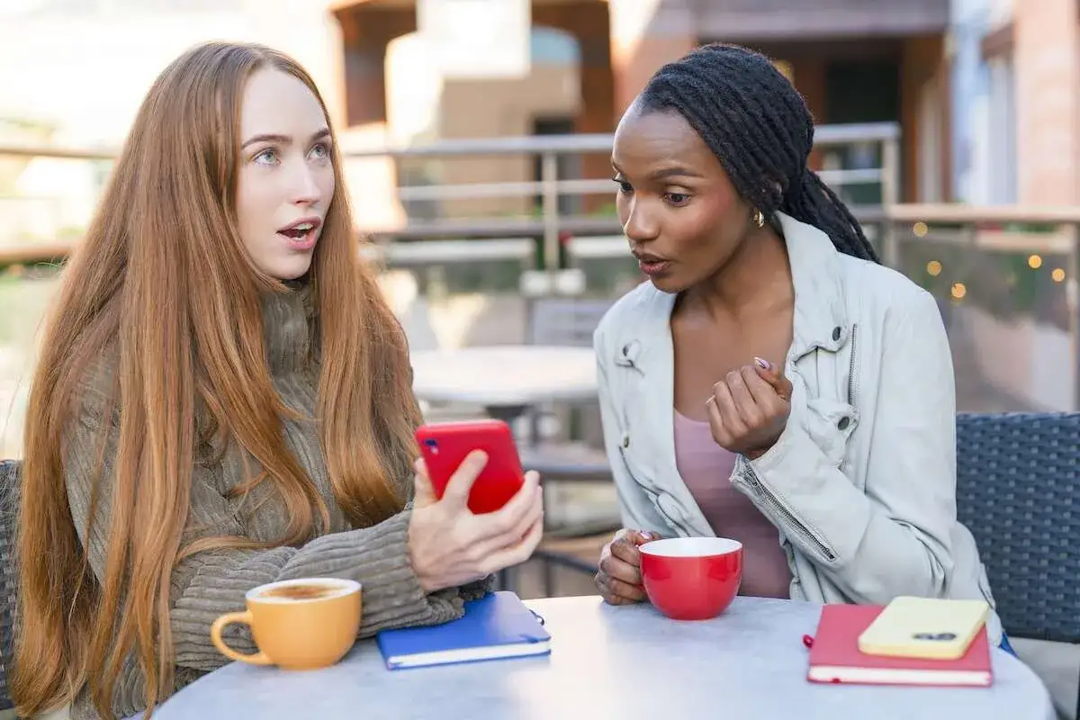Due amiche adulte si guardano sorprese mentre guardano un telefono rosso, con caffè e libri sul tavolo.
