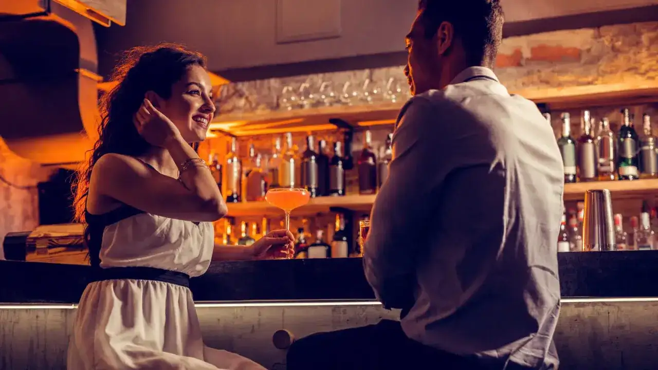 Una donna sorride mentre un uomo la guarda negli occhi da lontano, entrambi seduti al bancone di un bar con cocktail e bottiglie sullo sfondo.