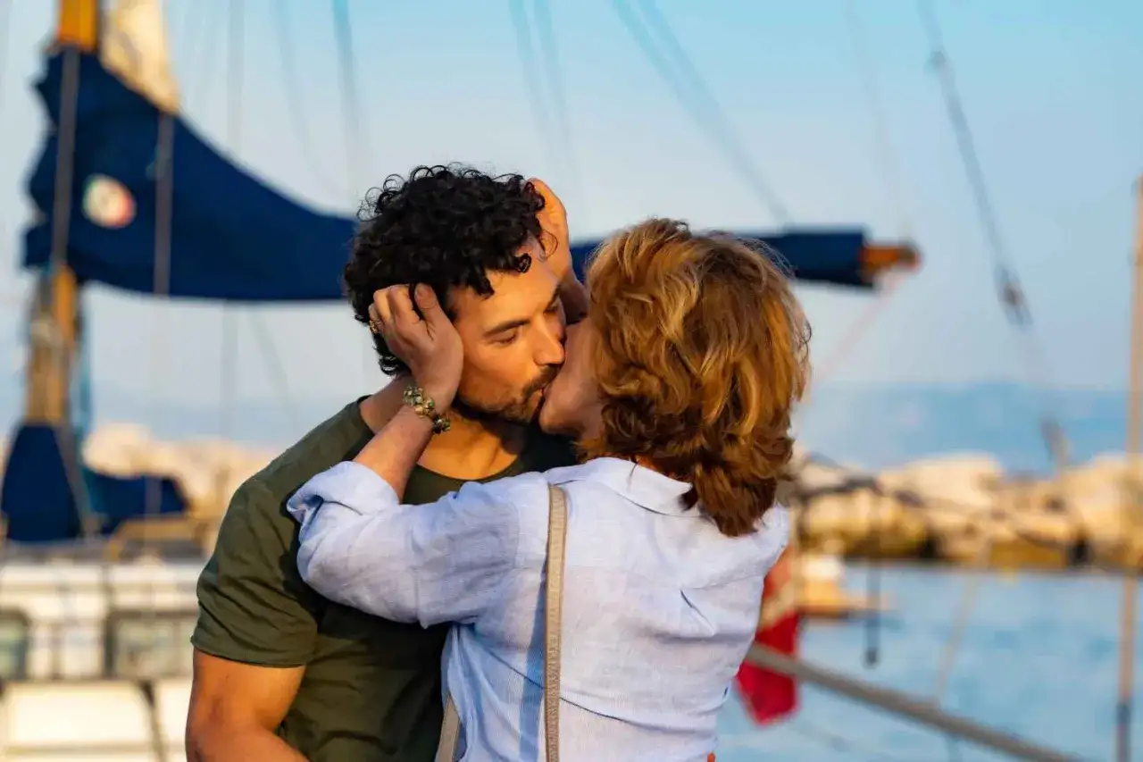 Un giovane uomo e una donna matura si baciano appassionatamente in un porto italiano, con barche a vela sullo sfondo.
