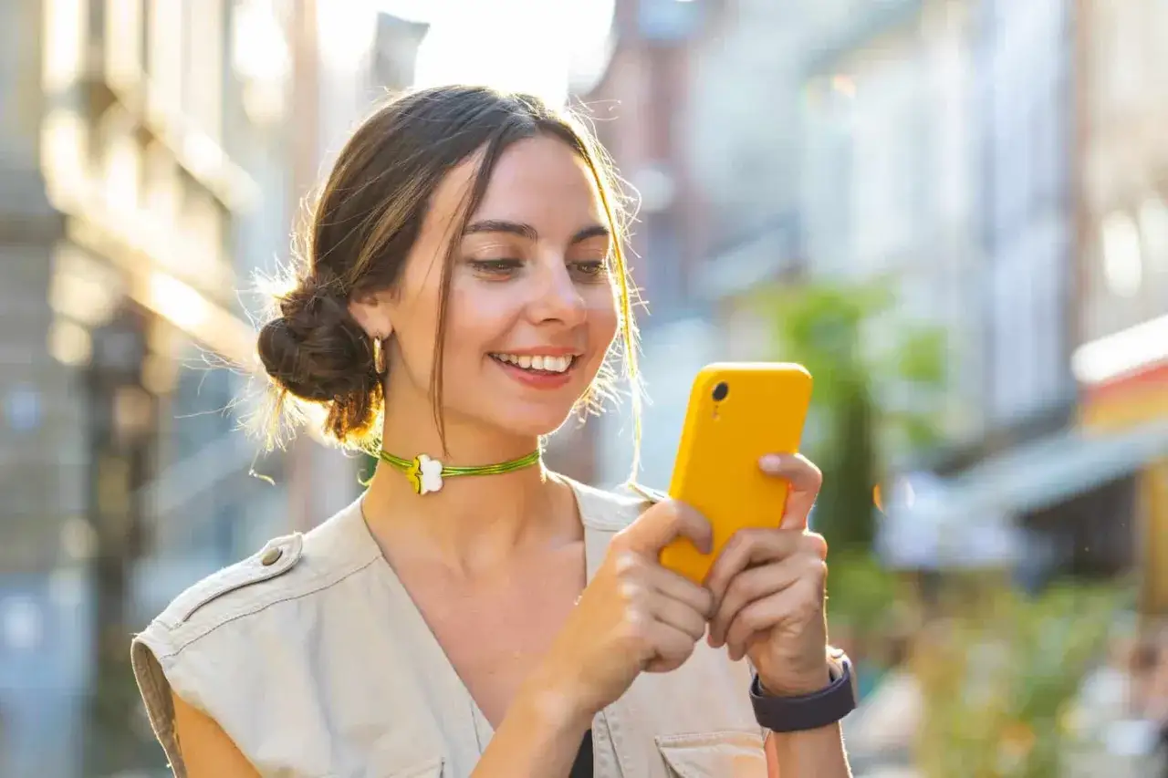 Ragazza sorridente con un telefono giallo, forse non sa cosa rispondere a un messaggio. Luce solare sui capelli raccolti.