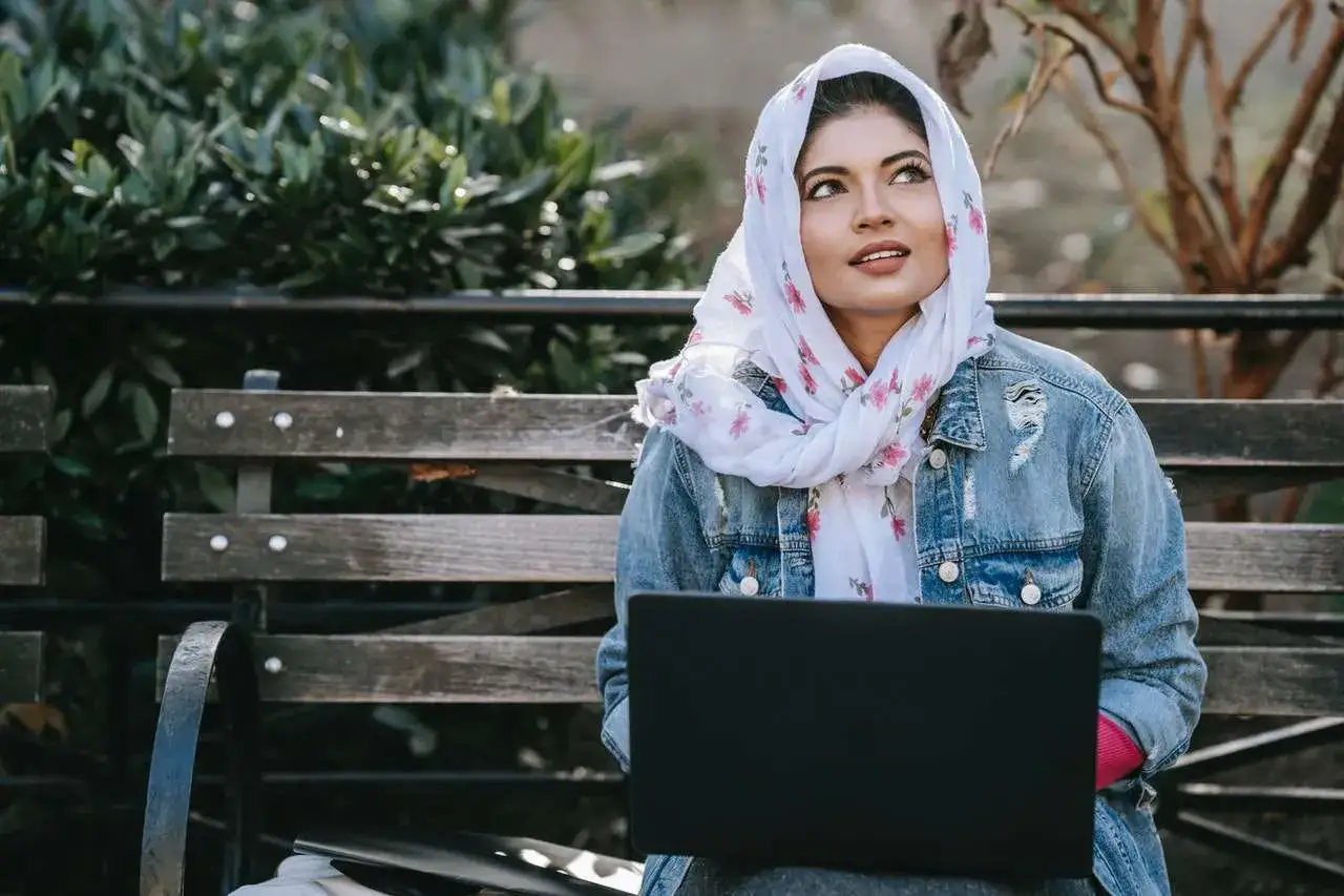 Ragazza con hijab floreale e giacca di jeans, laptop aperto, pensa se scrivere a un ragazzo o aspettare.