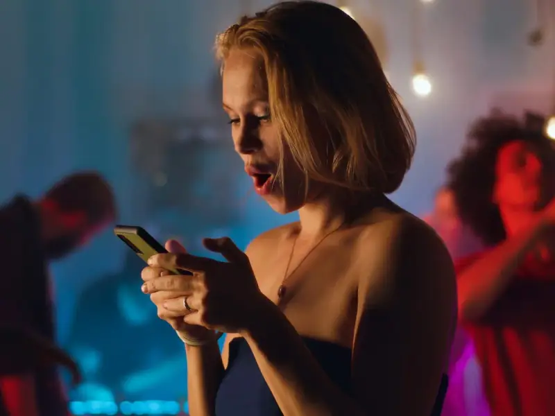 Sorpresa in chat: una ragazza con la bocca aperta guarda il suo telefono, mentre sullo sfondo si balla. Verità da fare in chat.